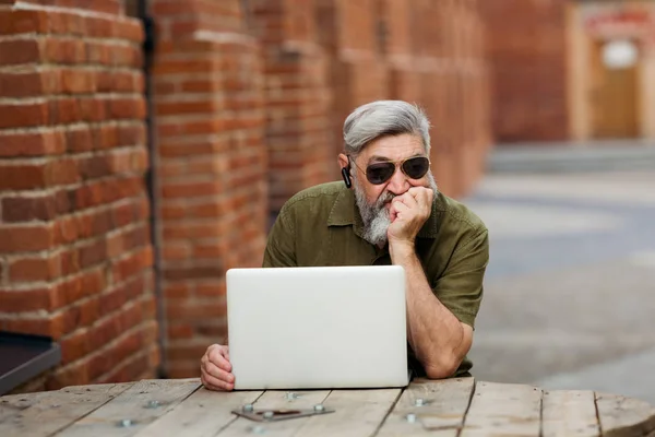 Gray adult businessman hipster in casual clothes sitting on the street near the office and unhappy and tired of working at a laptop