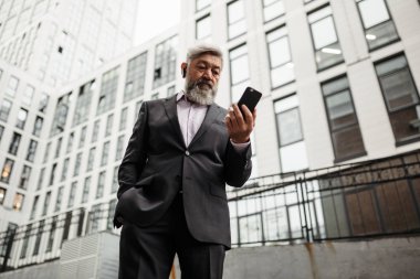 Takım elbiseli yaşlı sakallı bir adam bir iş bürosunun arka planında duruyor ve akıllı telefonun ekranına bakıyor.