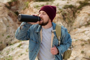 The traveler with a backpack on his shoulder quenches his thirst from a black thermos