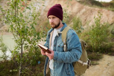 Writer in a hat with a book in his hands looking for inspiration on the shore of a blue mountain lake