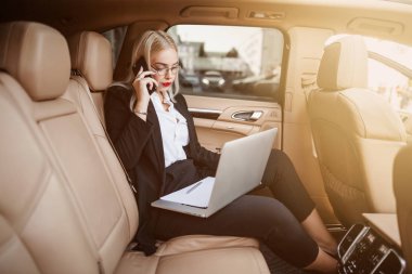 A businesswoman with blonde hair sits in the back of an expensive car and draws up a contract on a laptop