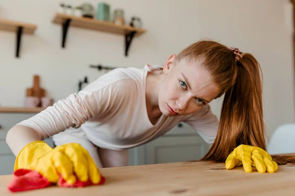A red-haired girl in yellow rubber gloves carefully cleans the kitchen table before starting to clean the house.