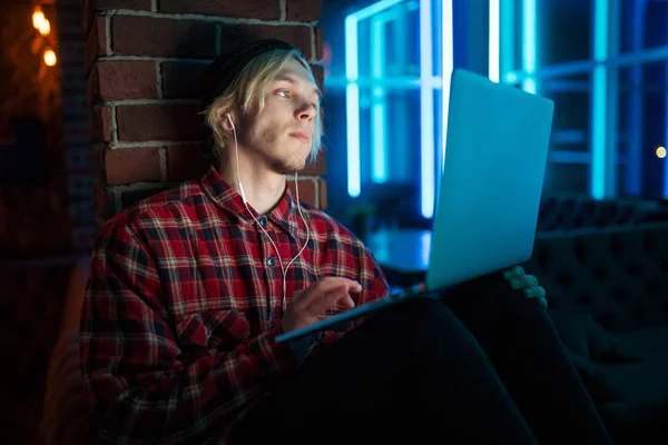 A hipster man listens to music with headphones and works on a laptop in a dark room of the bar against the background of blue neon signs