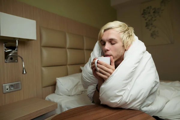 A young blond guy sitting on the bed holding a white mug in his hands and drinking hot coffee wrapped in a white blanket