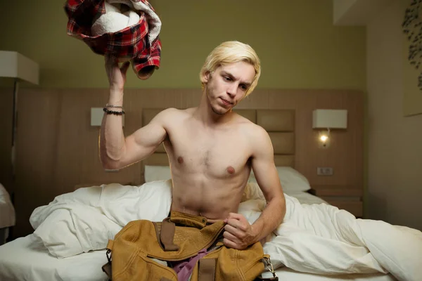 The blond guy sits on a bed with a naked torso in a hotel room and throws his things out of the suitcase with clothes