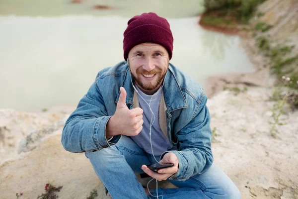 A bearded hipster tourist in a denim hoodie sits on the shore of a lake and listens to music through smartphone headphones. Shows like and looks at the camera