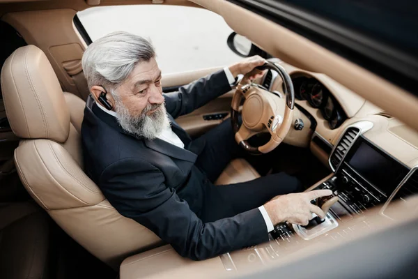 A gray bearded hipster businessman in a business suit sits behind the wheel of an expensive business class car