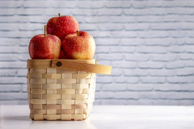 Elmalar ahşap sepetin üzerinde beyaz bir masada beyaz tuğlalı bir duvarın üzerinde. 