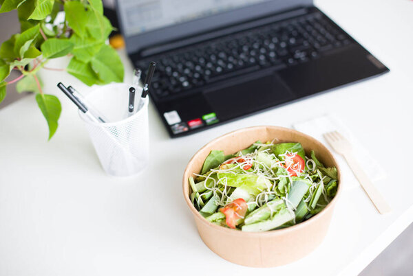 A fresh delivered salad bowl placed on an office desk next to a laptop, concept of convenient food delivery and healthy eating. High-quality photo.