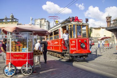 İstanbul, Türkiye - 05 Ağustos 2019: Ünlü İstiklal Caddesi'nin Taksim ucunda geleneksel Türk simiti satan bir arabanın yanında nostaljik tramvay. Turistler ve yerliler güneşin tadını çıkarıyor.