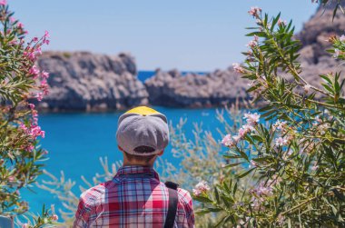 İsimsiz bir adam güneşli bir yaz gününde etrafı ağaçlarla çevrili mavi denize bakıyor. Seyahat eden mutlu bir turist..