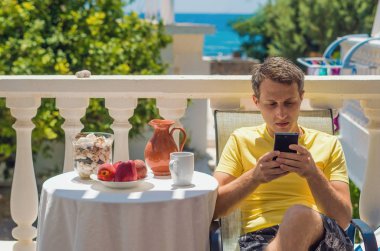 Adam cep telefonunu deniz kenarında güneşli bir terasta otururken kullanıyor. Mükemmel bir sabah haberleri ve sağlıklı kahvaltıların tadını çıkarıyorum..