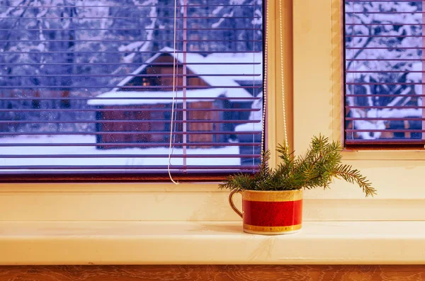 Cozy interior with a fir tree branch in a cup standing on a window sill near the window leading into snowy yard near the forest in the countryside