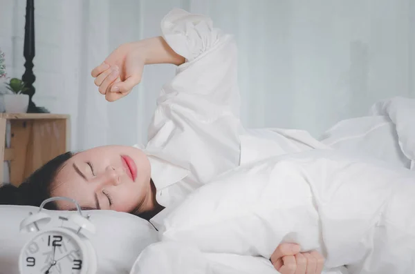 Sleepy asian young woman in bed with eyes closed extending hand to alarm clock at home.