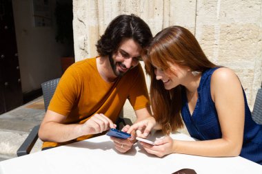 Genç çift bir yaz terasında cep bakıyor, onlar beklerken gülümseme