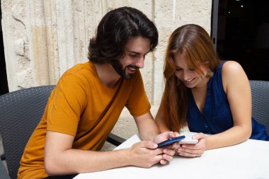 Genç çift bir yaz terasında cep bakıyor, onlar beklerken gülümseme