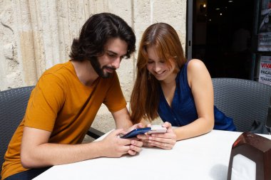 Genç çift bir yaz terasında cep bakıyor, onlar beklerken gülümseme