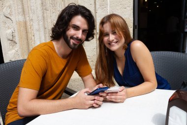 Genç çift bir yaz terasında cep bakıyor, kameraya bakarak gülümseme