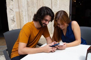 Genç çift bir yaz terasında cep bakıyor, onlar beklerken gülümseme