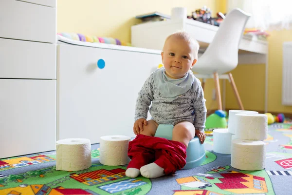 Smiling baby sitting on chamber pot with lots of toys and toilet paper around him in kids room