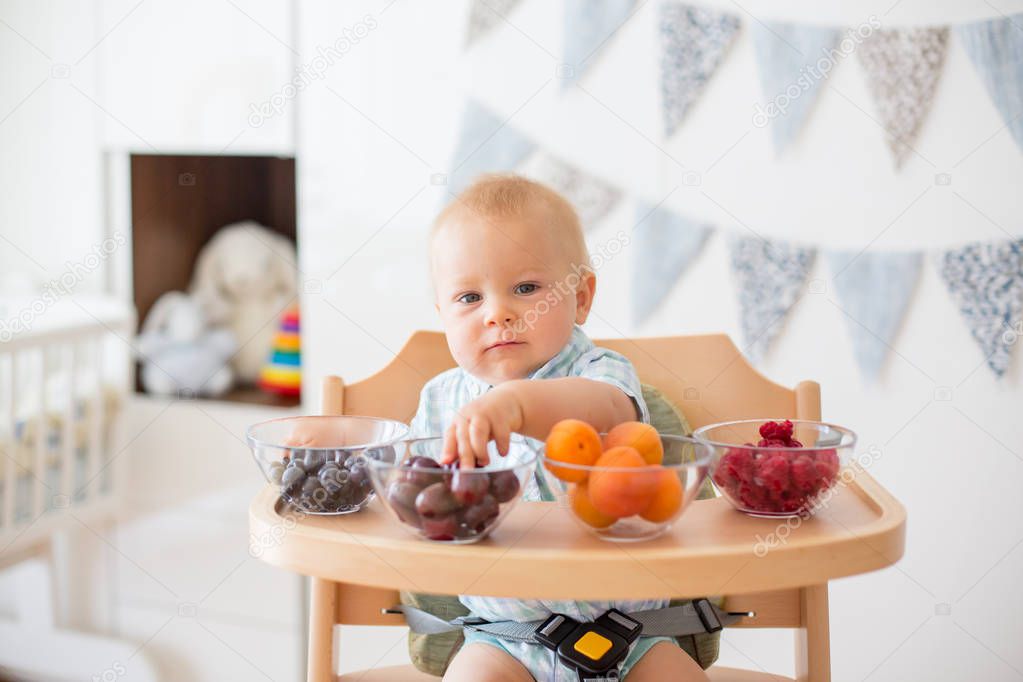 Adorable little baby boy, eating fresh fruits at home, sitting in baby chair in kids room