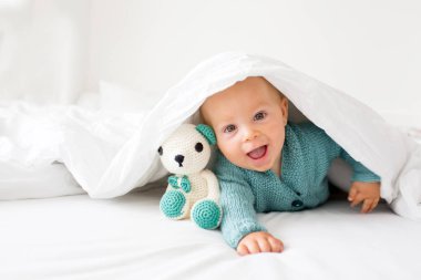 Mutlu bir şekilde beyaz güneşli, parlak yatak odasında kamera gülümseyen tutmak örme oyuncak, küçük şirin bebek çocuk, çocuk örme kazaklı,