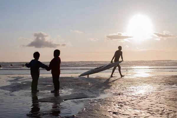 两个孩子的剪影 看冲浪者在海滩 日落时间 — 图库照片