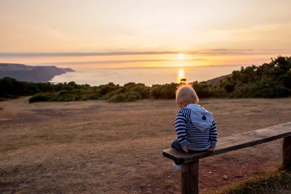 美しい赤ちゃん男の子 ベンチに座っている夏に海の上すばらしい夕日を考えて — ストック写真