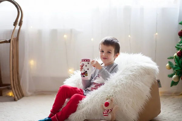 かわいい男の子 豆袋の上に座って お茶を飲んで クリスマス休暇を楽しんで彼の周りのクリスマスの装飾 — ストック写真