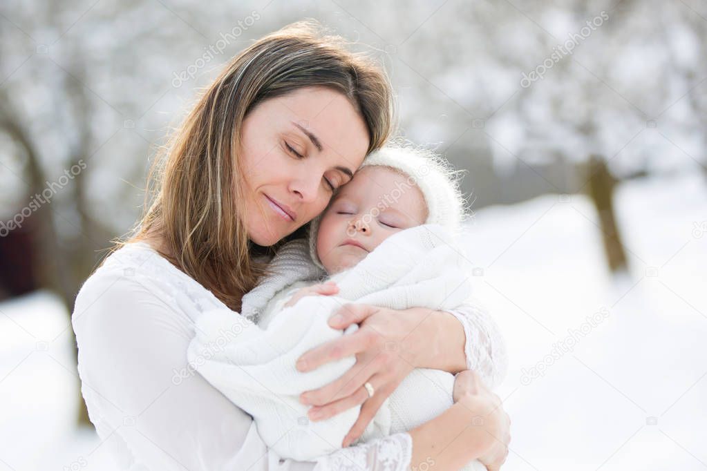 Beautiful mother in white dress and cute baby boy in knitted onesie, having taken their beautiful winter outdoor portrait on a sunny winter snowy day