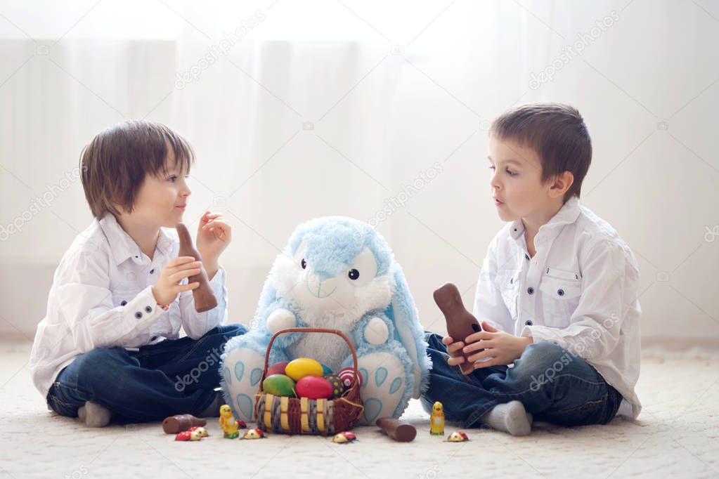Two adorable little children, boy brothers, having fun eating chocolate bunnies and playing with eggs at home, back light