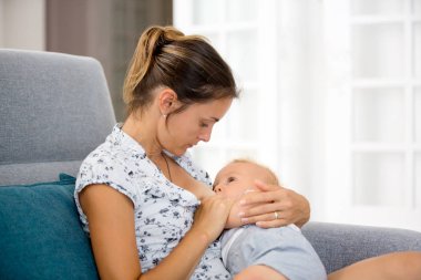 Onun toddler bebek çocuk, emzirme onu şefkatle sarılma güneşli oturma odasında evde oturan genç anne