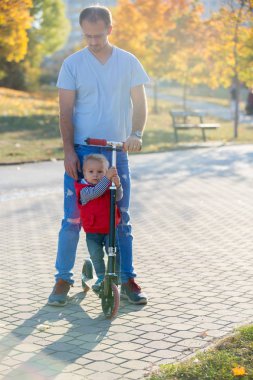 Babası onun bir sonbahar parkta yürümeye başlayan çocuk evlat scooter sürme. . Etkin aile boş. Spor, boş zaman çocuklar konsepti ile.