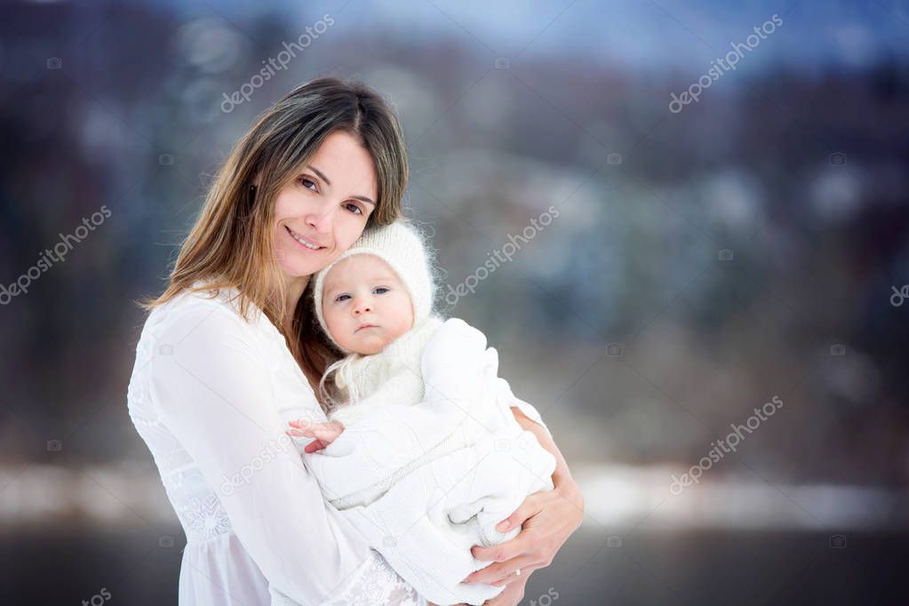 Beautiful mother in white dress and cute baby boy in knitted onesie, having taken their beautiful winter outdoor portrait on a sunny winter snowy day