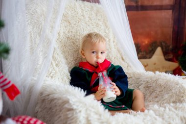 Noel ağacı ve arka planda oda ışıkları ile sallanan sandalyede oturup, uyku, kurabiye yemek Noel Baba bornoz giymiş erkek bebek, sevimli çocuk. Mutlu Noeller. Tatil sezonu.