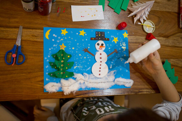 Top view of child, doing snowman and winter tree. Scissors, glue stick, pencil, markers, paper sheets and scraps on wooden background. Fun children winter background