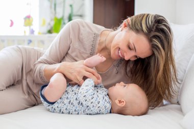 Genç anne, onun yeni doğan bebek çocuk ile yatakta yalan, oyun ve birlikte etkileşim. Aile mutluluk kavramı. Arka ışık fron pencerenin arkasında