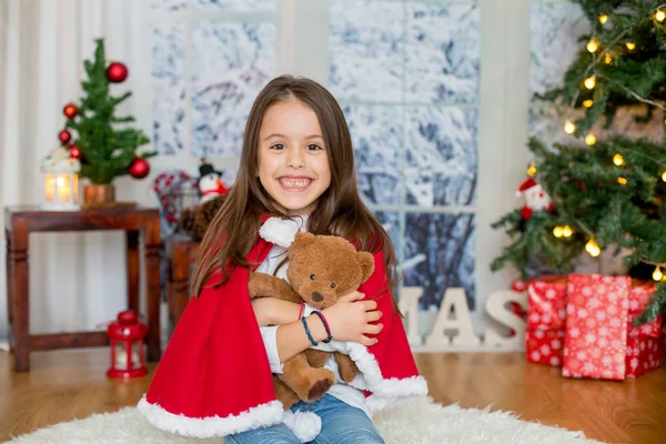 クリスマス ツリーの前に座っておもちゃで幸せな子 子供はサンタ クロースのドレスに身を包んだ 家庭で楽しい女の子 — ストック写真