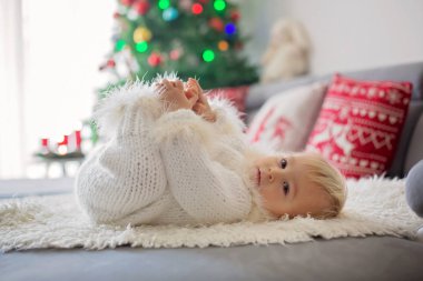 El örgüsü beyazı giyinmiş, kanepeye uzanmış hediyelerle oynayan ve mutlu bir şekilde gülümseyen meraklı küçük çocuk.