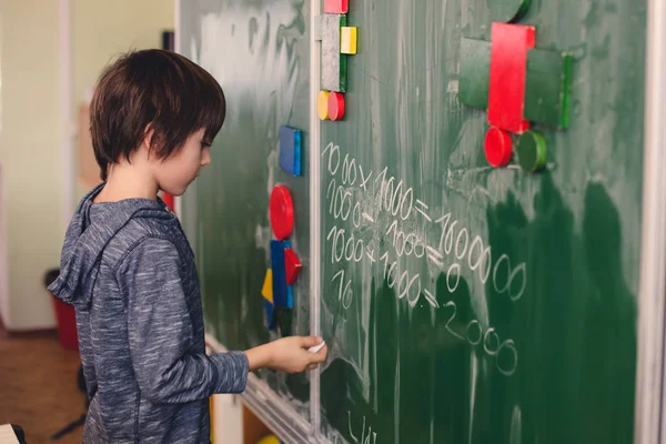 First grade child, learning math, shapes and colors at school, standing in front of blackboard