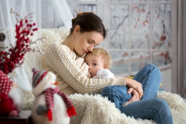Anne, bebeğini emziriyor rahat koltukta oturuyor, kış vakti kar yağıyor.