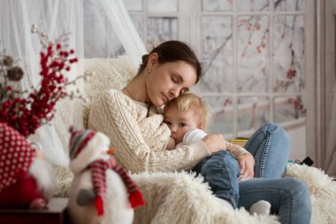 Anne, bebeğini emziriyor rahat koltukta oturuyor, kış vakti kar yağıyor.