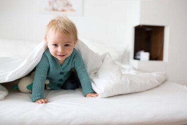 Mutlu bir şekilde beyaz güneşli, parlak yatak odasında kamera gülümseyen tutmak örme oyuncak, küçük şirin bebek çocuk, çocuk örme kazaklı,