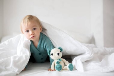 Mutlu bir şekilde beyaz güneşli, parlak yatak odasında kamera gülümseyen tutmak örme oyuncak, küçük şirin bebek çocuk, çocuk örme kazaklı,