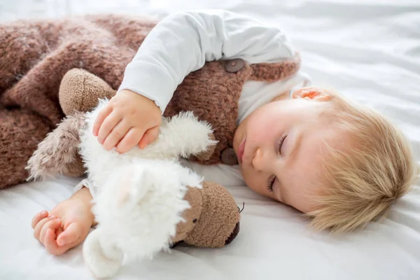 Sweet baby boy in cute overall, sleeping in bed with teddy bear stuffed toys, winter landscape behind him