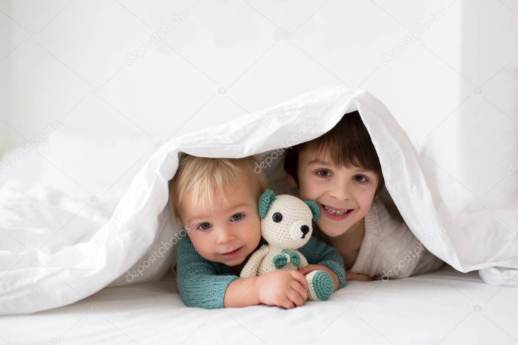 Little cute baby boy and his older brother, children in knitted sweaters, holding knitted toy, smiling happily at camera in white sunny, bright bedroom