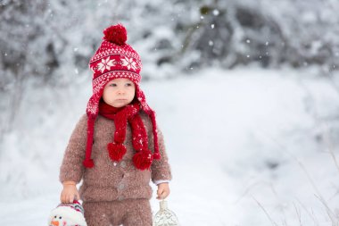 Fener ve kardan adam, ceket, pantolon, kırmızı şapka ve atkı, örgü kahverengi bir elinde giyinmiş sevimli bebek bebek portresi zevk ilk kar karlı parkta yürüyor