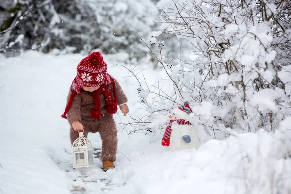 ランタンや雪だるま ニット ジャケット ズボン 赤い帽子とスカーフ 褐色の手に身を包んだかわいい幼児赤ちゃんの肖像画を吹く最初の雪を楽しむ雪の公園を歩く — ストック写真