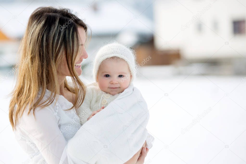 Beautiful mother in white dress and cute baby boy in knitted onesie, having taken their beautiful winter outdoor portrait on a sunny winter snowy day