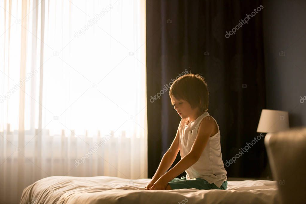 Two sibling children, baby boy and his older brother, sitting on bed in sunny bedroom, playing together, back light